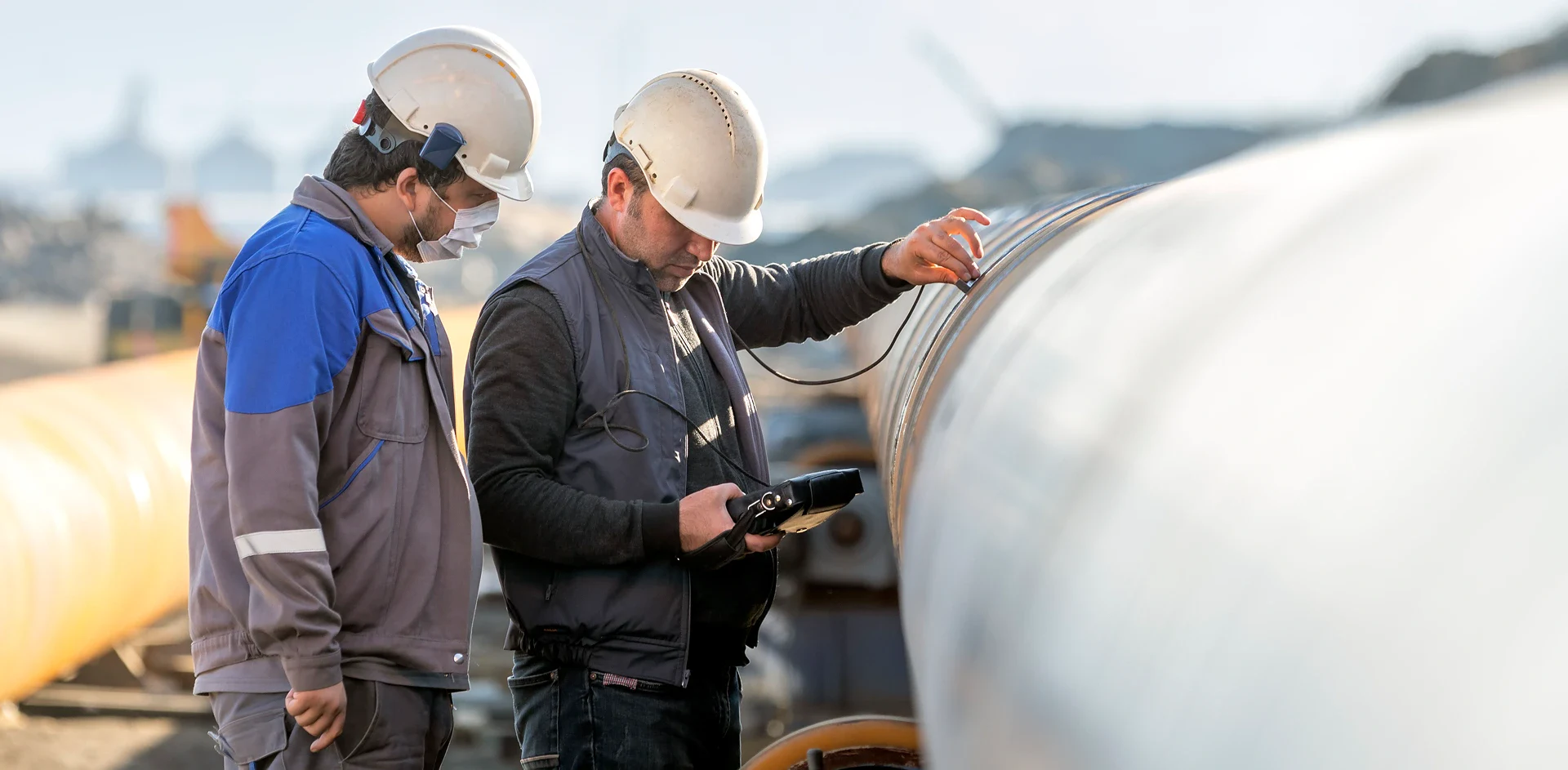 Two workers in safety gear inspect a large pipe, using a device to conduct measurements. The setting is an industrial site.