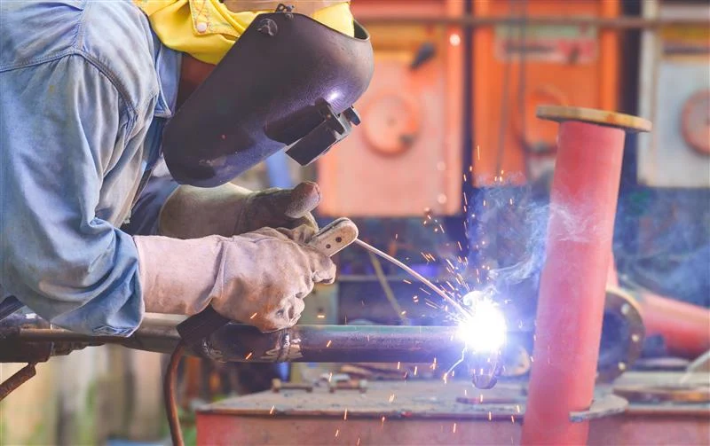 A worker in a bright yellow rain jacket and hard hat kneels by a ship's hull, taking notes on a clipboard near the water.