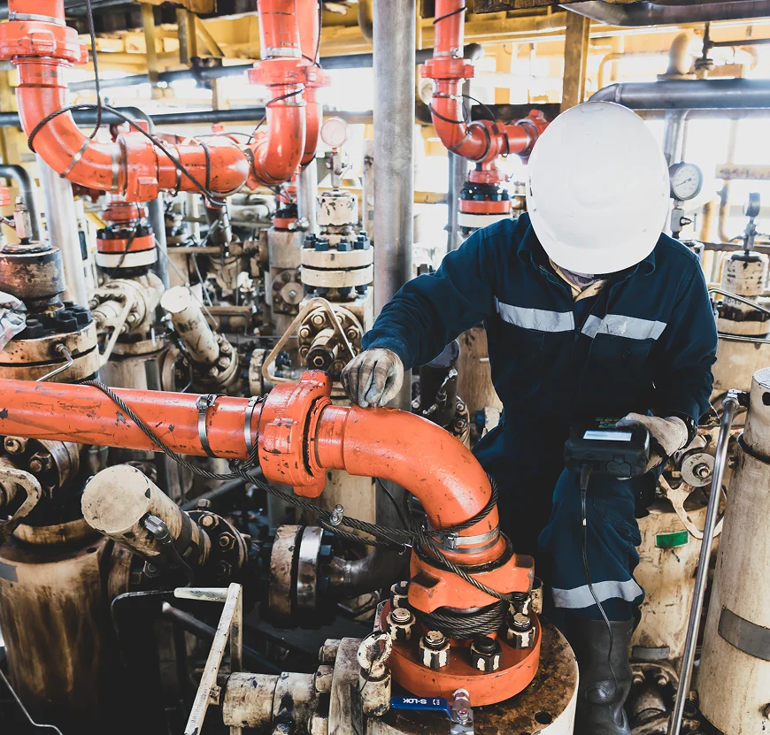 A worker in a hard hat is inspecting orange pipes and valves in an industrial setting, using a handheld device to monitor equipment.