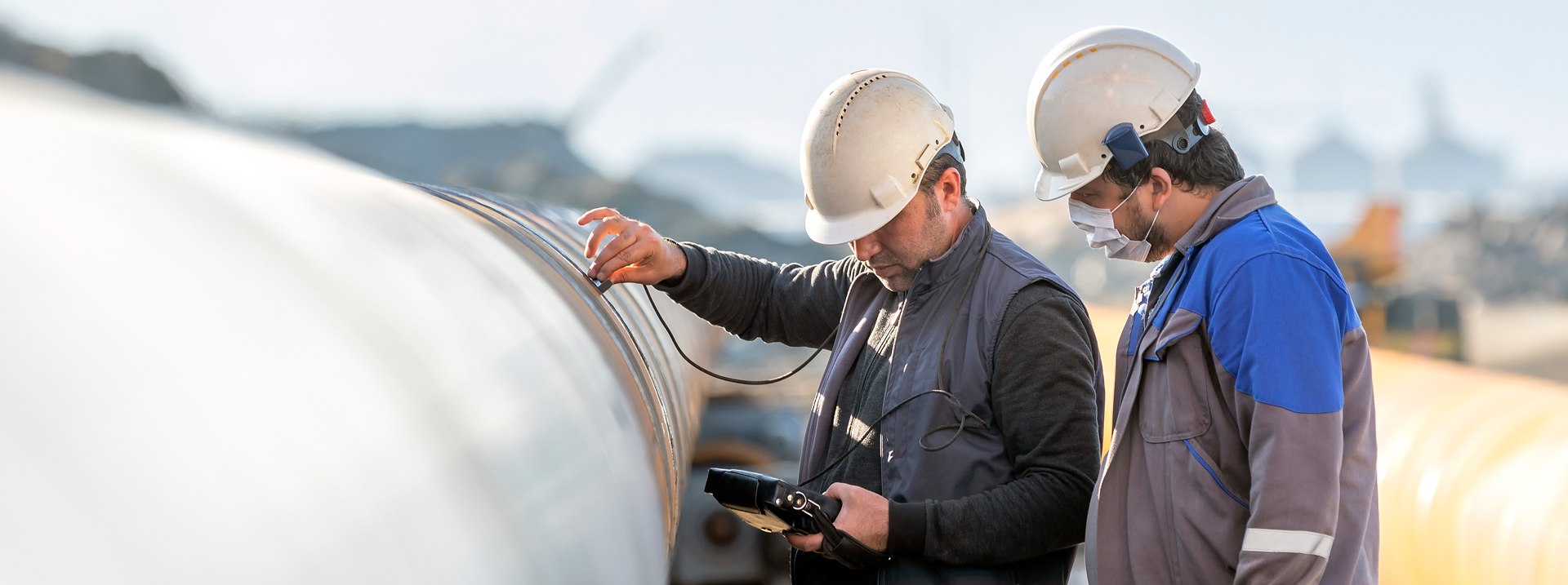 Two workers in helmets inspect a large pipe, one using a testing device, against an industrial background.
