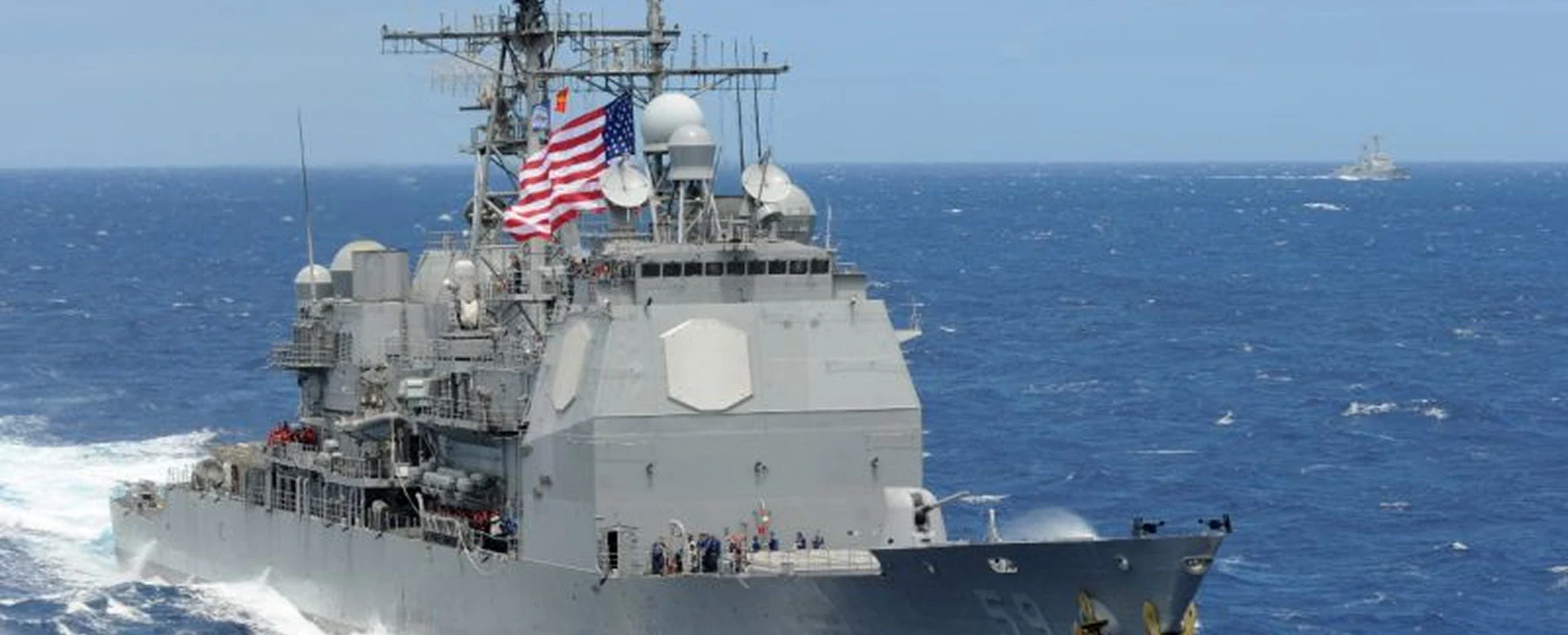 A naval ship navigates through ocean waves, showcasing its sleek gray hull and equipment against a backdrop of blue water.